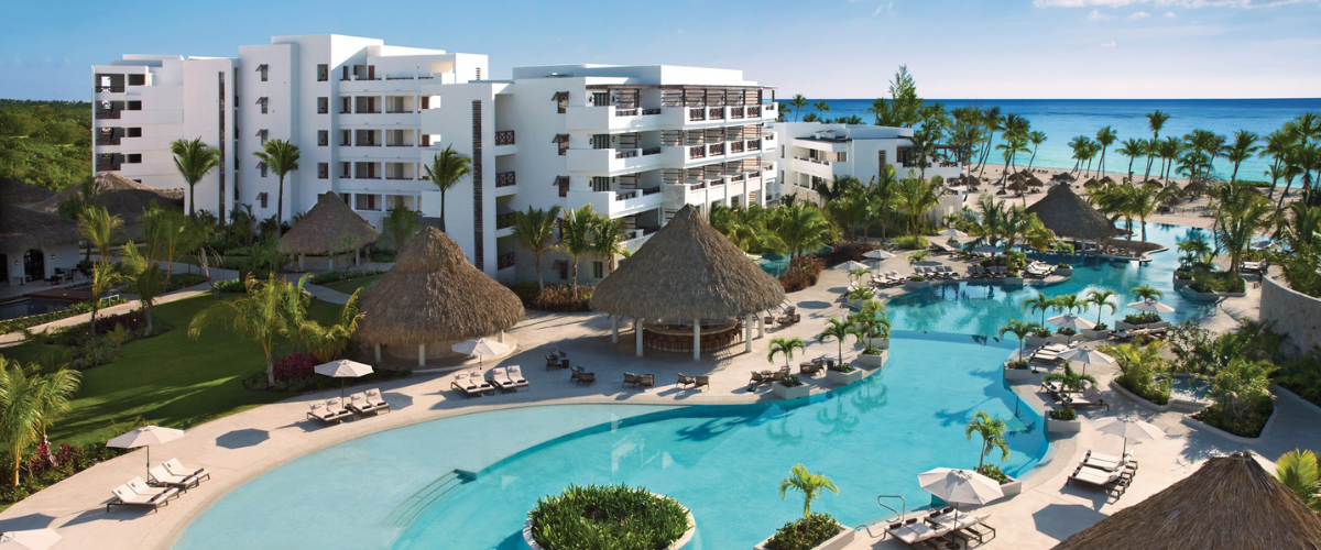 Aerial view of an all-inclusive resort showing the buildings with a large pool surrounded by palm trees and the ocean in the background 