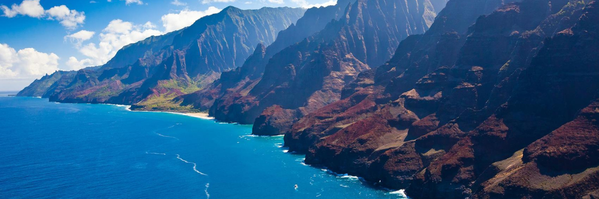 Aerial view of a mountain in Hawaii with water at the foot of the mountain with blue skies and white clouds behind