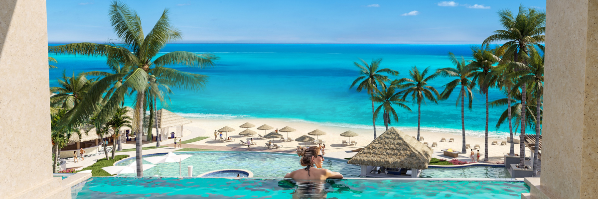 View from an in room plunge pool with a woman leaning against the side of the pool looking over the resort at the main pool and sandy beach with the ocean in the background and palm trees around