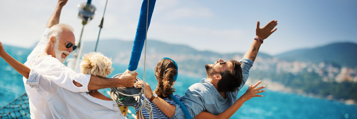 Close up of four people hanging on to a post of the edge of a sailboat on the water with water and land view in the background