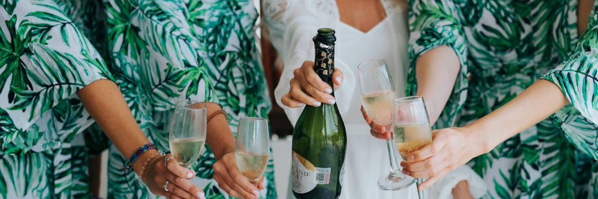 Close up of four bridesmaids in white robes with green ferns and a bride with a white robe holding up a champagne bottle while the bridesmaids hold up champagne glasses 