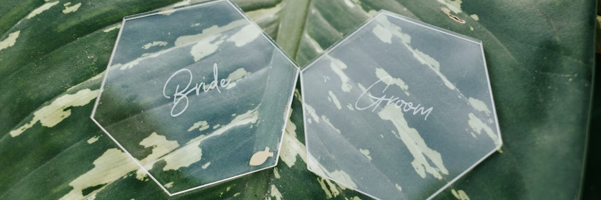 Two clear hexagon plates engraved with bride and groom on them laying on top of a big green leaf