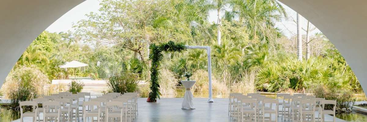 Destination wedding set up under a covering with white chairs and a simple arch with green leafy arrangement around half of it