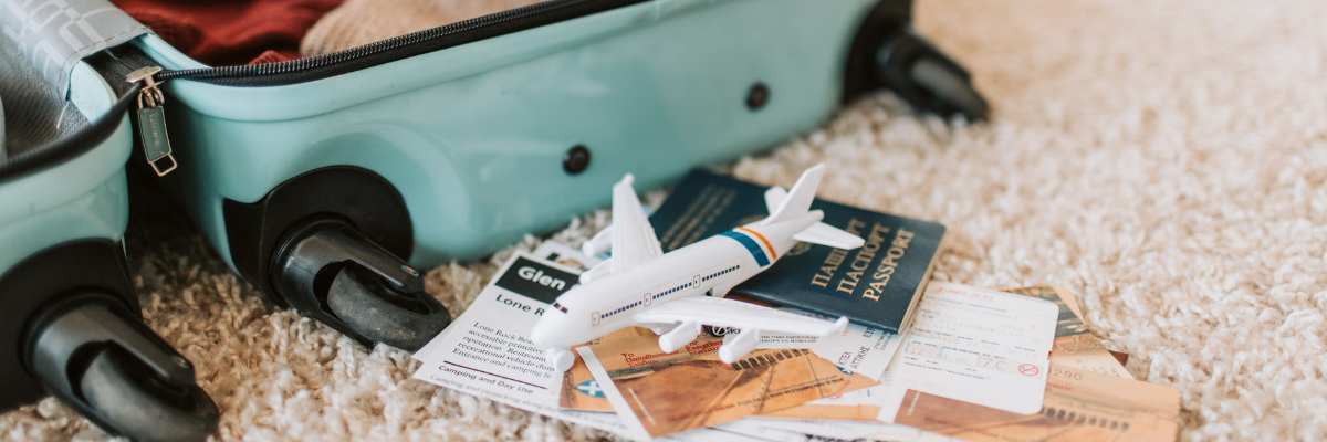 A blue suitcase laid open on a white carpet with clothes packed in side of it and a small toy plane next to it with a passport and travel papers underneath.