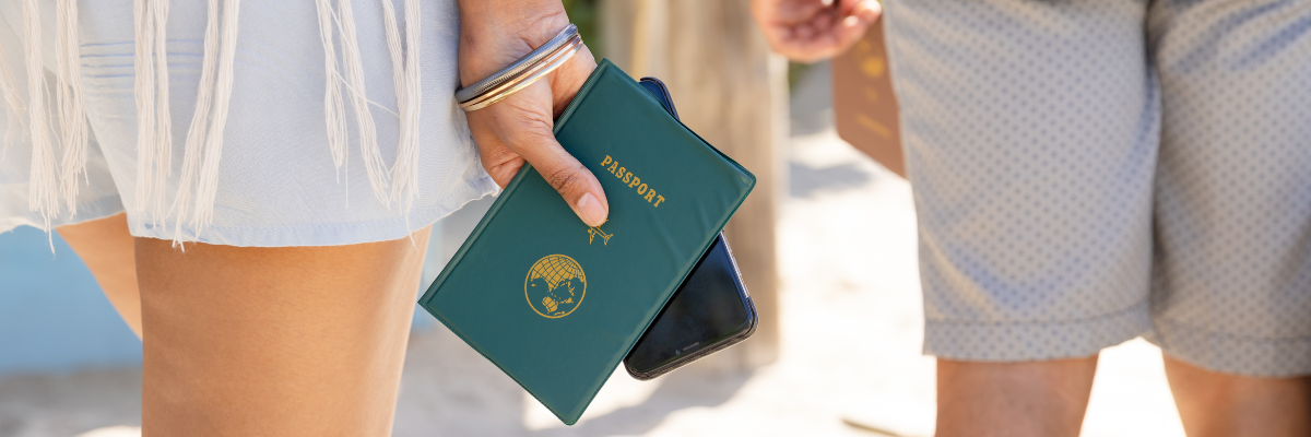 A close up of a ladies hand holding a green passport and a phone with a guy standing slightly in front of her with a brown passport in his hand.